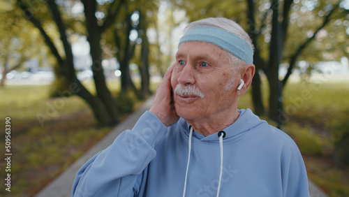 Portrait of athletic fitness senior elderly sport runner man training, wearing earphones listening music, looking at camera and smiling. Healthy grandfather workout cardio outdoors in park at morning