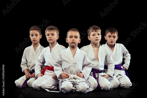 Group portrait of preschool age boys, beginner karate fighters in white doboks posing like team isolated on dark background. Concept of sport, martial arts, education