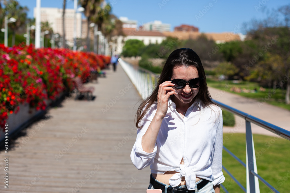 Young millennial woman walking on the bridge with colorful red flowers enjoying the sun and life, smiling happily