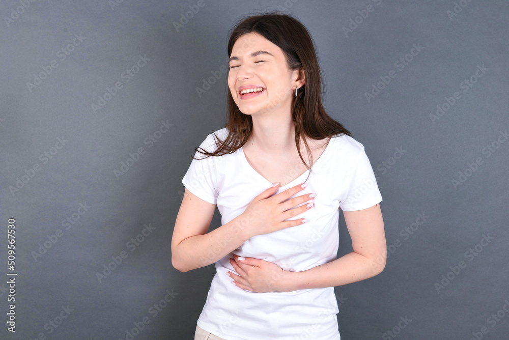 young beautiful Caucasian woman wearing white T-shirt over grey wall keeps hands crossed, laughs out loud at good joke, wears casual clothes. young beautiful Caucasian woman wearing white T-shirt over grey wall keeps hands crossed, laughs out loud at good joke, wears casual clothes.
