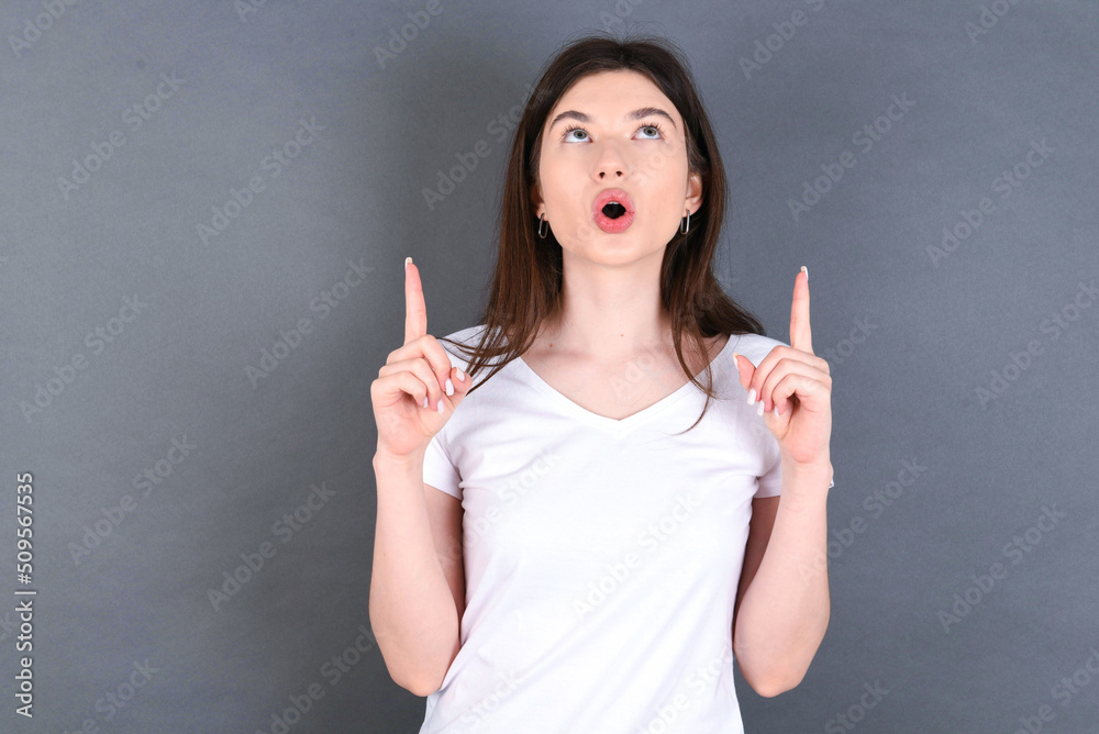 young beautiful Caucasian woman wearing white T-shirt over grey wall being amazed and surprised looking and pointing up with fingers showing something strange.
