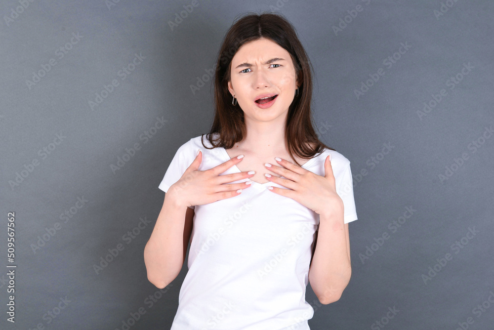 young beautiful Caucasian woman wearing white T-shirt over grey wall keeps hands on chest feeling shocked and scared, mouth widely opened, stares at camera saying: Who, me? young beautiful Caucasian woman wearing white T-shirt over grey wall keeps hands on chest feeling shocked and scared, mouth widely opened, stares at camera saying: Who, me?