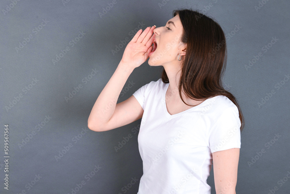 young beautiful Caucasian woman wearing white T-shirt over grey wall profile view, looking happy and excited, shouting and calling to copy space. young beautiful Caucasian woman wearing white T-shirt over grey wall profile view, looking happy and excited, shouting and calling to copy space.