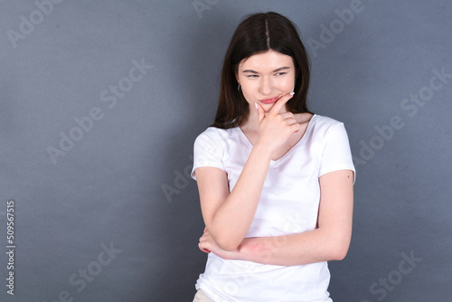 Thoughtful smiling young beautiful Caucasian woman wearing white T-shirt over grey wall keeps hand under chin, looks directly at camera, listens something with interest. Youth concept.