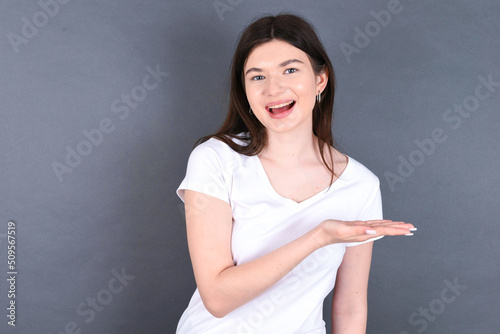 young beautiful Caucasian woman wearing white T-shirt over grey wall says: wow how exciting it is, has amazed expression, shows something on blank space with palm. Advertisement concept.