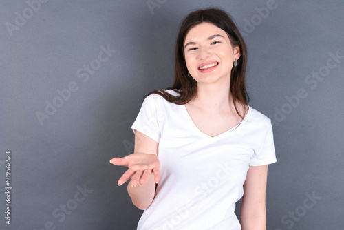 young beautiful Caucasian woman wearing white T-shirt over grey wall smiling friendly offering something with open hand or handshake as greeting and welcoming. Successful business.