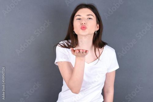 young beautiful Caucasian woman wearing white T-shirt over grey wall looking at the camera blowing a kiss with hand on air being lovely. Love expression.