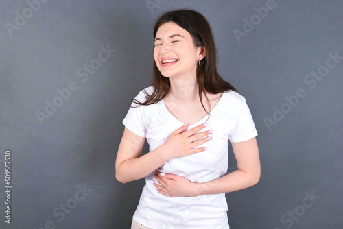 young beautiful Caucasian woman wearing white T-shirt over grey wall keeps hands crossed, laughs out loud at good joke, wears casual clothes.