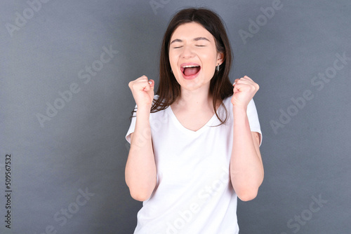 young beautiful Caucasian woman wearing white T-shirt over grey wall rejoicing his success and victory clenching fists with joy being happy to achieve aim and goals. Positive emotions, feelings.