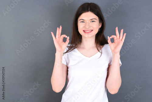 Glad young beautiful Caucasian woman wearing white T-shirt over grey wall shows ok sign with both hands as expresses approval, has cheerful expression, being optimistic.