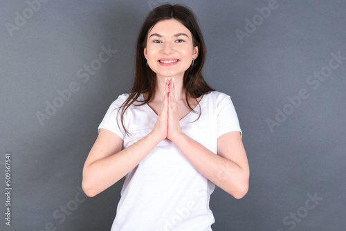 young beautiful Caucasian woman wearing white T-shirt over grey wall keeps palms together, has pleased expression. Glad attractive male makes request, pleads for mercy. Hopeful young adult.