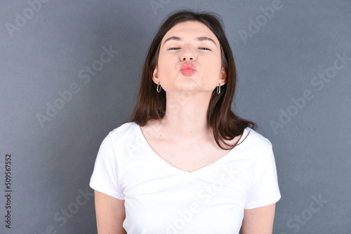young beautiful Caucasian woman wearing white T-shirt over grey wall, keeps lips as going to kiss someone, has glad expression, grimace face. Standing indoors. Beauty concept.