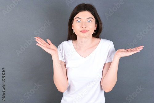 Puzzled and clueless young beautiful Caucasian woman wearing white T-shirt over grey wall with arms out  shrugging shoulders  saying  who cares  so what  I don t know.