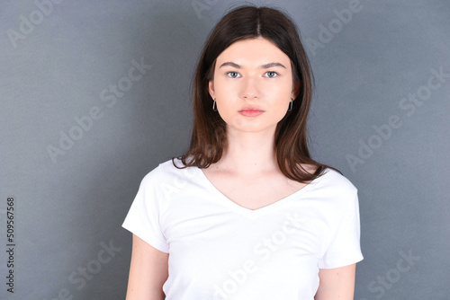 Joyful young beautiful Caucasian woman wearing white T-shirt over grey wall looking to the camera, thinking about something. Both arms down, neutral facial expression.