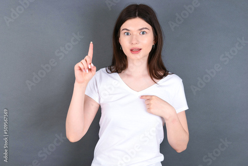 young beautiful Caucasian woman wearing white T-shirt over studio grey says: wow how exciting it is, has amazed expression, indicates something. One hand on her chest and pointing with other hand.
