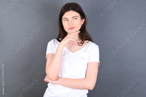 Dreamy young beautiful Caucasian woman wearing white T-shirt over studio grey wall with pleasant expression, looks sideways, keeps hand under chin, thinks about something pleasant.