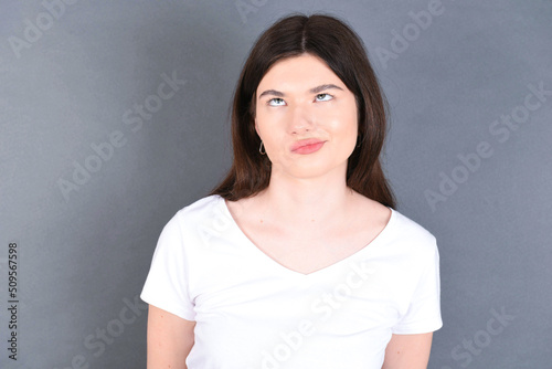 young beautiful Caucasian woman wearing white T-shirt over studio grey wall has worried face looking up lips together, being upset thinking about something important, keeps hands down.