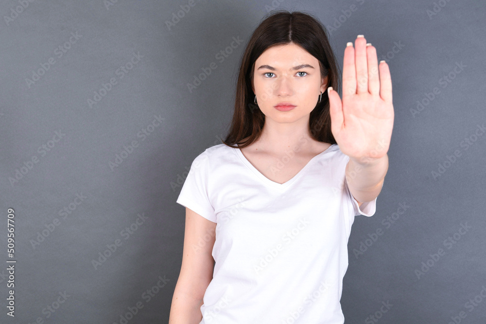 young beautiful Caucasian woman wearing white T-shirt over studio grey wall doing stop gesture with palm of the hand. Warning expression with negative and serious gesture on the face. young beautiful Caucasian woman wearing white T-shirt over studio grey wall doing stop gesture with palm of the hand. Warning expression with negative and serious gesture on the face.