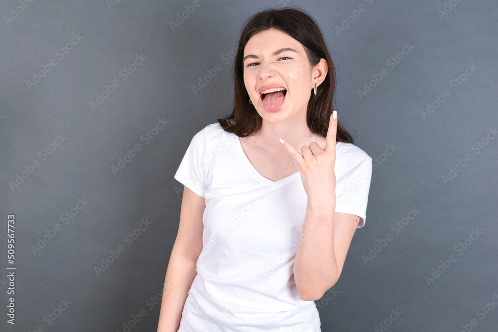 Portrait of a crazy young beautiful Caucasian woman wearing white T-shirt over studio grey wall showing tongue horns up gesture, expressing excitement of being on concert of band. Portrait of a crazy young beautiful Caucasian woman wearing white T-shirt over studio grey wall showing tongue horns up gesture, expressing excitement of being on concert of band.