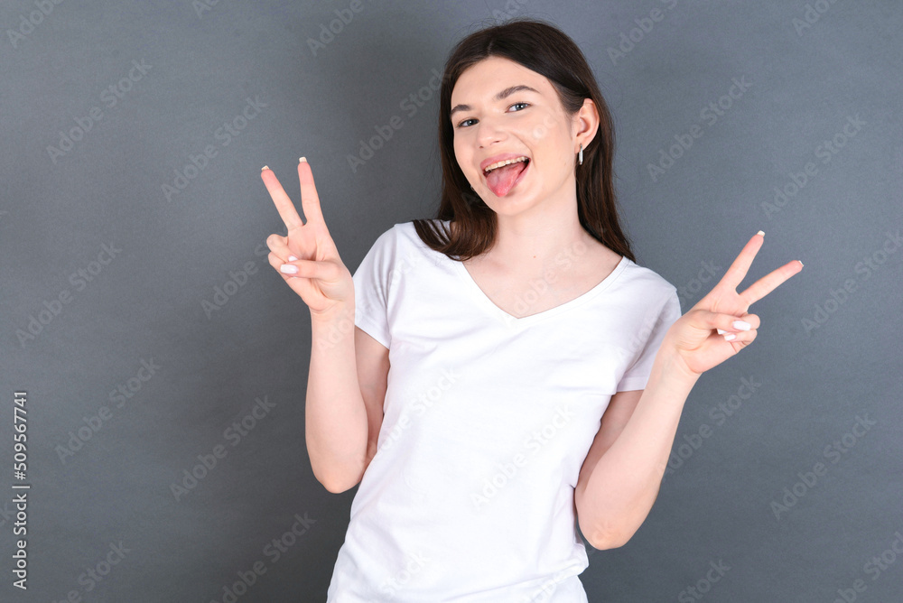 young beautiful Caucasian woman wearing white T-shirt over studio grey wall with optimistic smile, showing peace or victory gesture with both hands, looking friendly. V sign. young beautiful Caucasian woman wearing white T-shirt over studio grey wall with optimistic smile, showing peace or victory gesture with both hands, looking friendly. V sign.