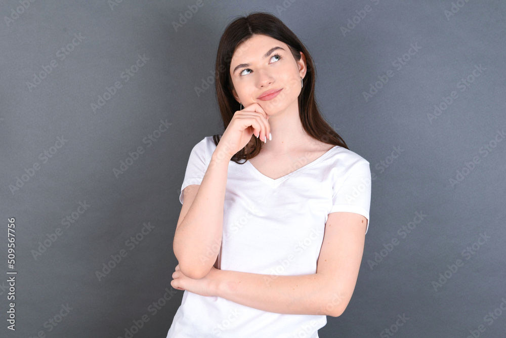Face expressions and emotions. Thoughtful young beautiful Caucasian woman wearing white T-shirt over studio grey wall holding hand under his head, having doubtful look.