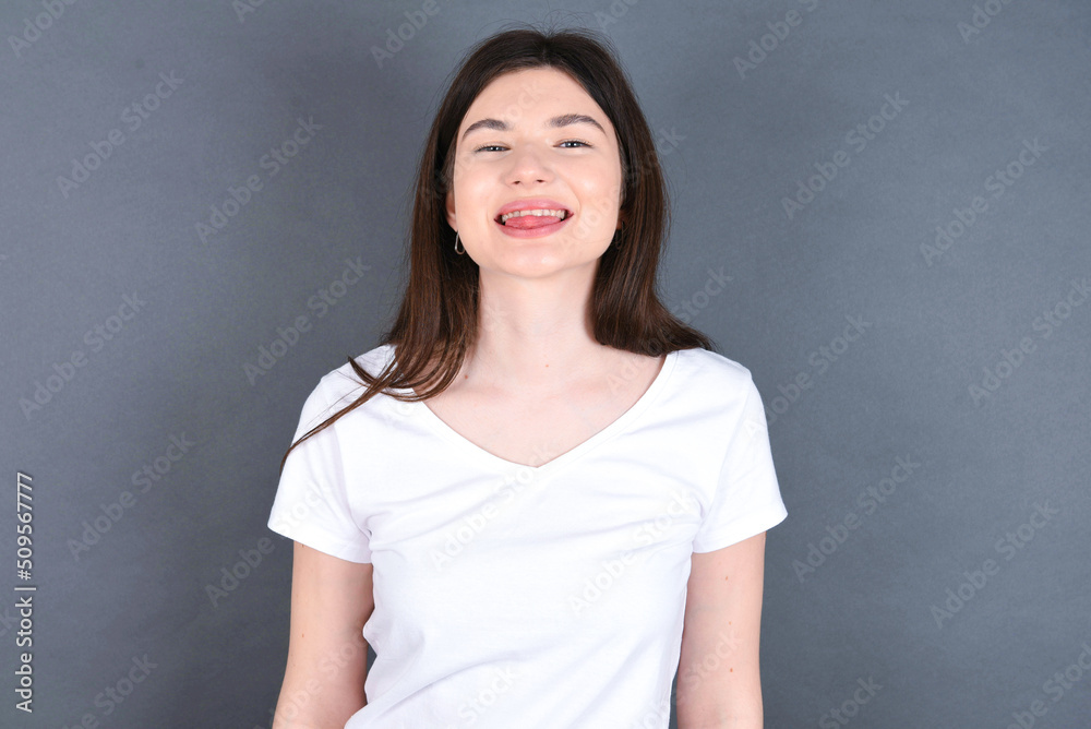 young beautiful Caucasian woman wearing white T-shirt over studio grey wall with happy and funny face smiling and showing tongue. young beautiful Caucasian woman wearing white T-shirt over studio grey wall with happy and funny face smiling and showing tongue.