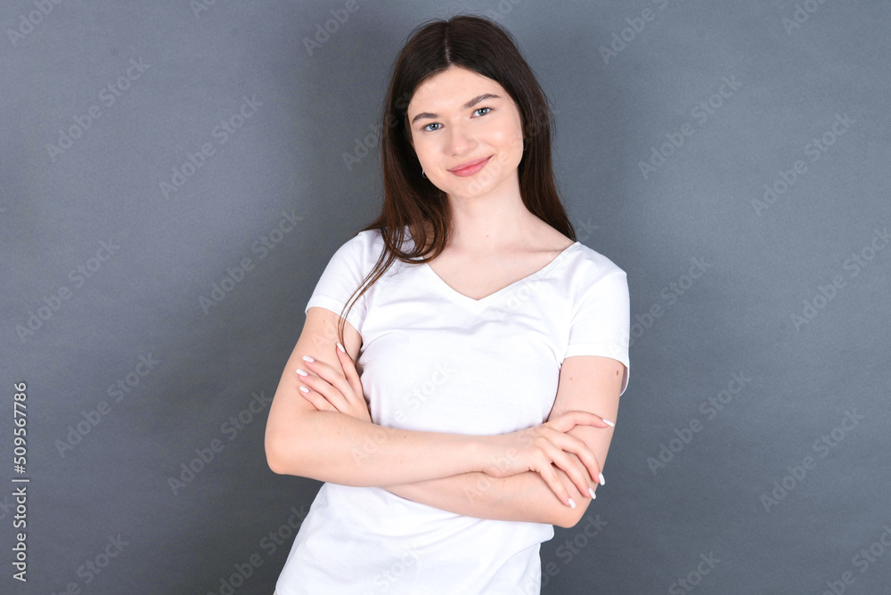 Waist up shot of self confident young beautiful Caucasian woman wearing white T-shirt over studio grey wall has broad smile, crosses arms, happy to meet with colleagues. Waist up shot of self confident young beautiful Caucasian woman wearing white T-shirt over studio grey wall has broad smile, crosses arms, happy to meet with colleagues.