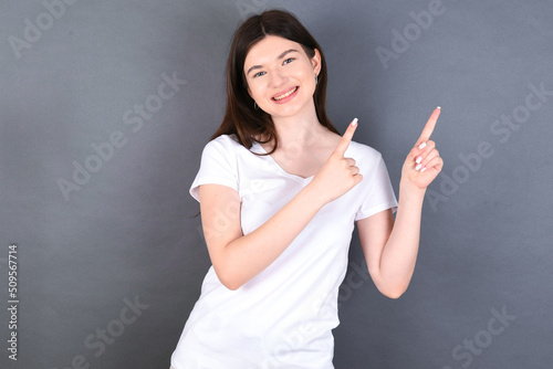 young beautiful Caucasian woman wearing white T-shirt over studio grey wall with positive expression, indicates with fore finger at blank copy space for your promotional text or advertisement.