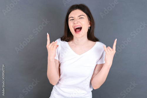 Born to rock this world. Joyful young beautiful Caucasian woman wearing white T-shirt over studio grey wall screaming out loud and showing with raised arms horns or rock gesture.