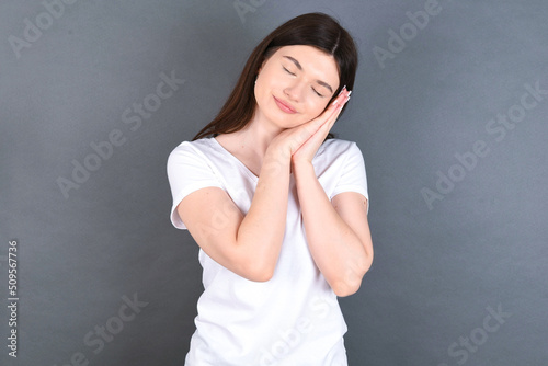 Relax and sleep time. Tired young beautiful Caucasian woman wearing white T-shirt over studio grey wall with closed eyes leaning on palms making sleeping gesture.