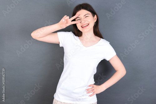 young beautiful Caucasian woman wearing white T-shirt over studio grey wall making v-sign near eyes. Leisure, coquettish, celebration, and flirt concept.