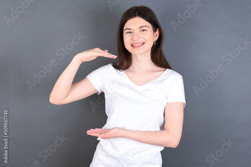 young beautiful Caucasian woman wearing white T-shirt over studio grey wall gesturing with hands showing big and large size sign, measure symbol.
