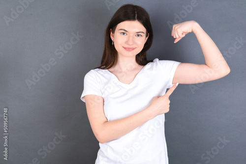 Smiling young beautiful Caucasian woman wearing white T-shirt over studio grey wall raises hand to show muscles, feels confident in victory, strong and independent.
