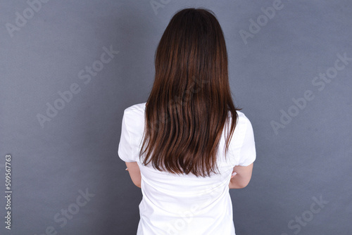 young beautiful Caucasian woman wearing white T-shirt over studio grey wall standing backwards looking away with arms on body.