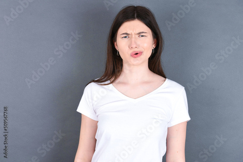 young beautiful Caucasian woman wearing white T-shirt over studio grey wall expressing disgust, unwillingness, disregard having tensive look frowning face, looking indignant with something.