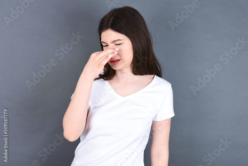young beautiful Caucasian woman wearing white T-shirt over studio grey wall, holding his nose because of a bad smell.