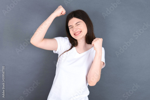 Attractive young beautiful Caucasian woman wearing white T-shirt over studio grey wall celebrating a victory punching the air with his fists and a beaming toothy smile.