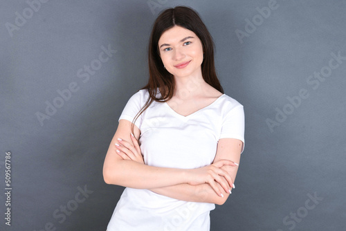 Waist up shot of self confident young beautiful Caucasian woman wearing white T-shirt over studio grey wall has broad smile, crosses arms, happy to meet with colleagues.