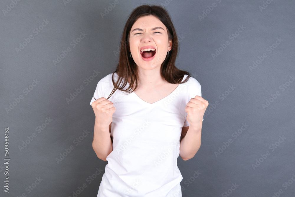 Joyful excited lucky young beautiful Caucasian woman wearing white T-shirt over studio grey wall cheering, celebrating success, screaming yes with clenched fists Joyful excited lucky young beautiful Caucasian woman wearing white T-shirt over studio grey wall cheering, celebrating success, screaming yes with clenched fists