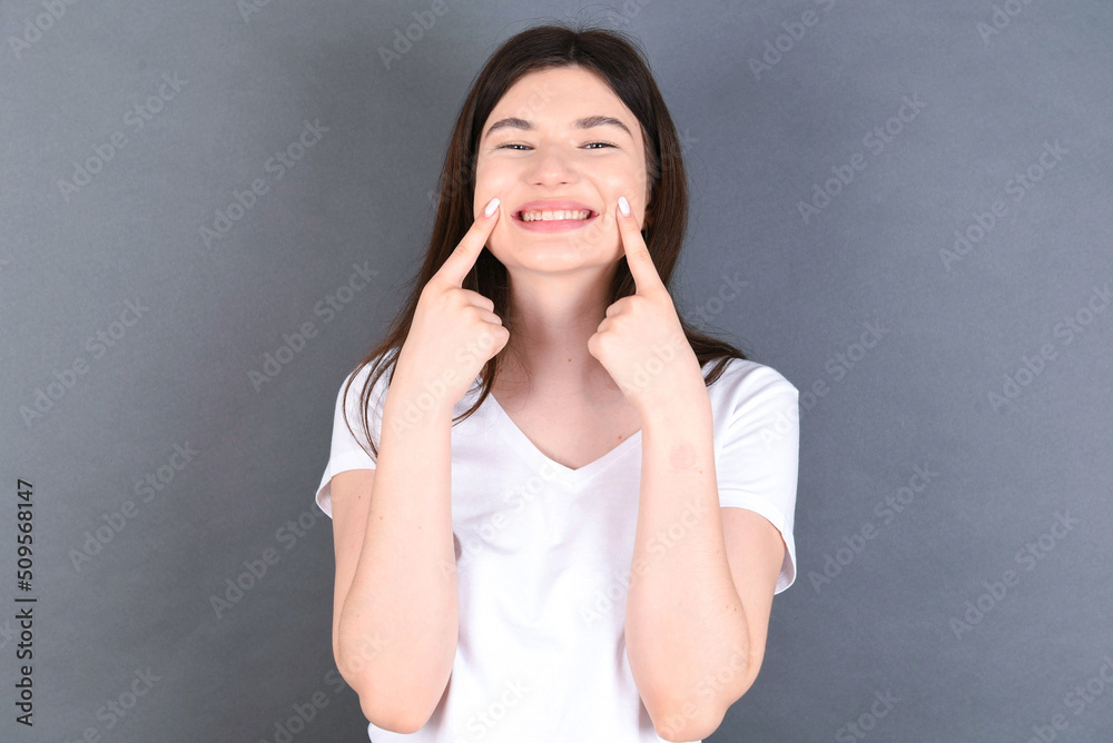 Strong healthy straight white teeth. Close up portrait of happy young beautiful Caucasian woman wearing white T-shirt over studio grey wall with beaming smile pointing on perfect clear white teeth. Strong healthy straight white teeth. Close up portrait of happy young beautiful Caucasian woman wearing white T-shirt over studio grey wall with beaming smile pointing on perfect clear white teeth.