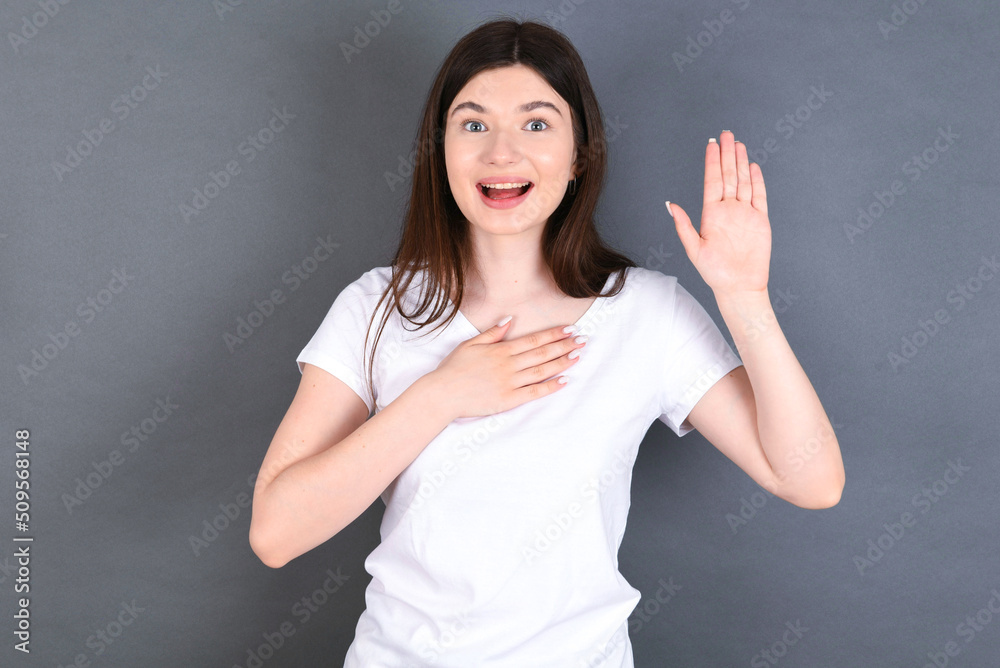 Fototapeta premium I swear, promise you not regret. Portrait of sincere young beautiful Caucasian woman wearing white T-shirt raising one arm and hold hand on heart as give oath, telling truth, want you to believe.