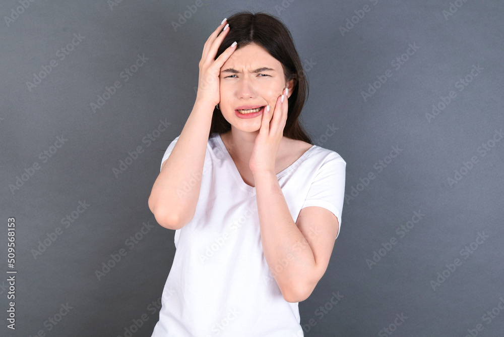 Young gloomy young beautiful Caucasian woman wearing white T-shirt over studio grey wall, hiding face with hands pouting and crying, standing upset and depressed complaining about job problem. Young gloomy young beautiful Caucasian woman wearing white T-shirt over studio grey wall, hiding face with hands pouting and crying, standing upset and depressed complaining about job problem.