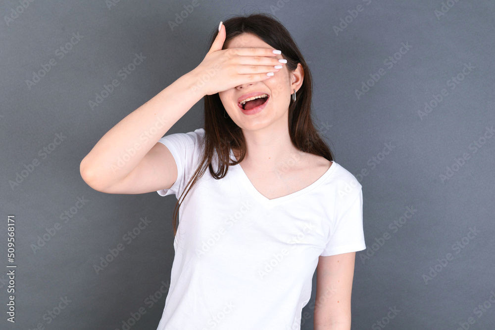Happy young beautiful Caucasian woman wearing white T-shirt over studio closing eyes with hand going to see surprise prepared by friend standing and smiling in anticipation for something wonderful. Happy young beautiful Caucasian woman wearing white T-shirt over studio closing eyes with hand going to see surprise prepared by friend standing and smiling in anticipation for something wonderful.
