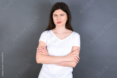young beautiful Caucasian woman wearing white T-shirt over studio grey wall frowning his face in displeasure, keeping arms folded, waiting for an explanation.