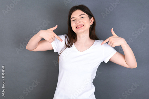 Pick me  Confident  self-assured and charismatic young beautiful Caucasian woman wearing white T-shirt over studio grey wall promoting oneself as wanting role smiling broadly and pointing at body.