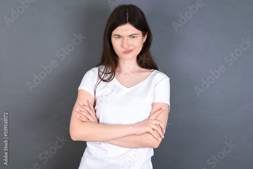 Picture of angry young beautiful Caucasian woman wearing white T-shirt over studio grey wall crossing arms. Looking at camera with disappointed expression.