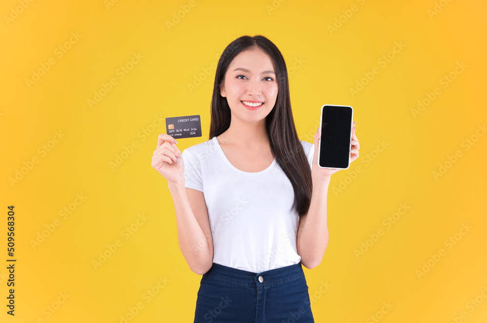 pretty asian woman smile showing, presenting credit card for payment or paying online in holding mobile phone payment credit card for confident money and financial concepts yellow background. pretty asian woman smile showing, presenting credit card for payment or paying online in holding mobile phone payment credit card for confident money and financial concepts yellow background.