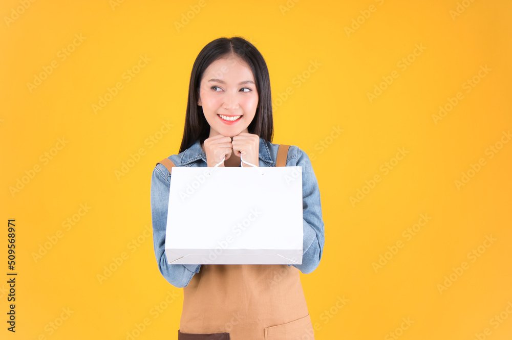 pretty asian female smile freelancer own shop show blank white paper bag to customers by products yellow background. pretty asian female smile freelancer own shop show blank white paper bag to customers by products yellow background.