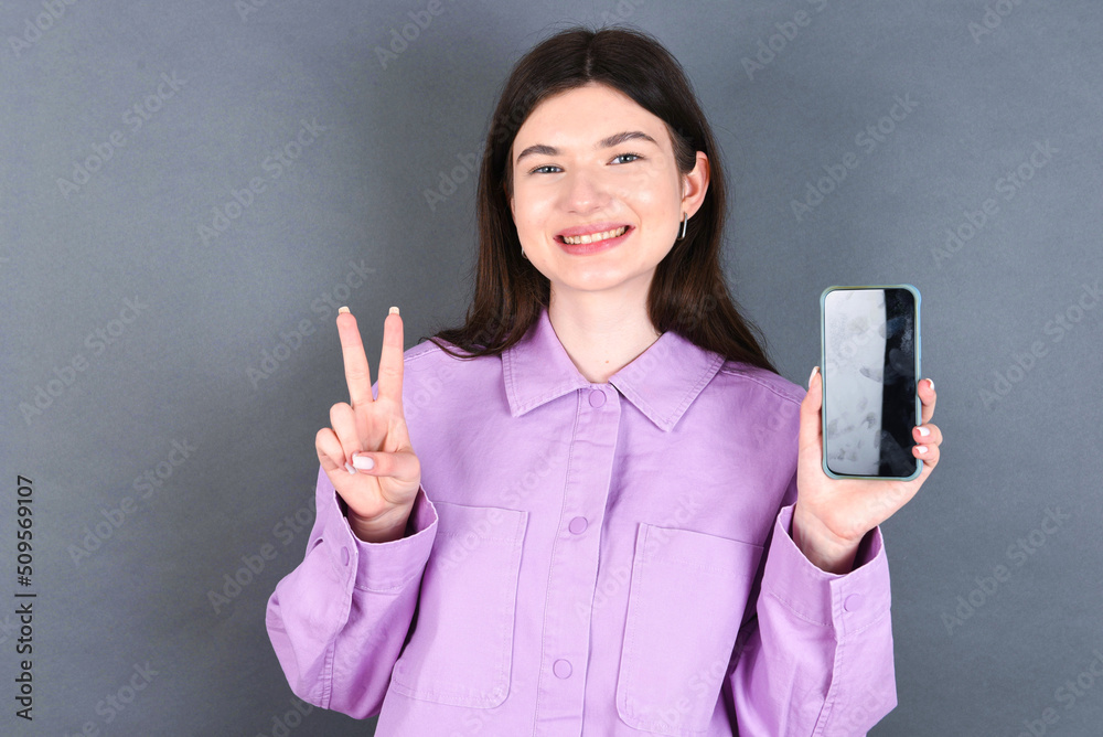 young beautiful Caucasian woman wearing pink shirt over studio grey wall holding modern device showing v-sign young beautiful Caucasian woman wearing pink shirt over studio grey wall holding modern device showing v-sign