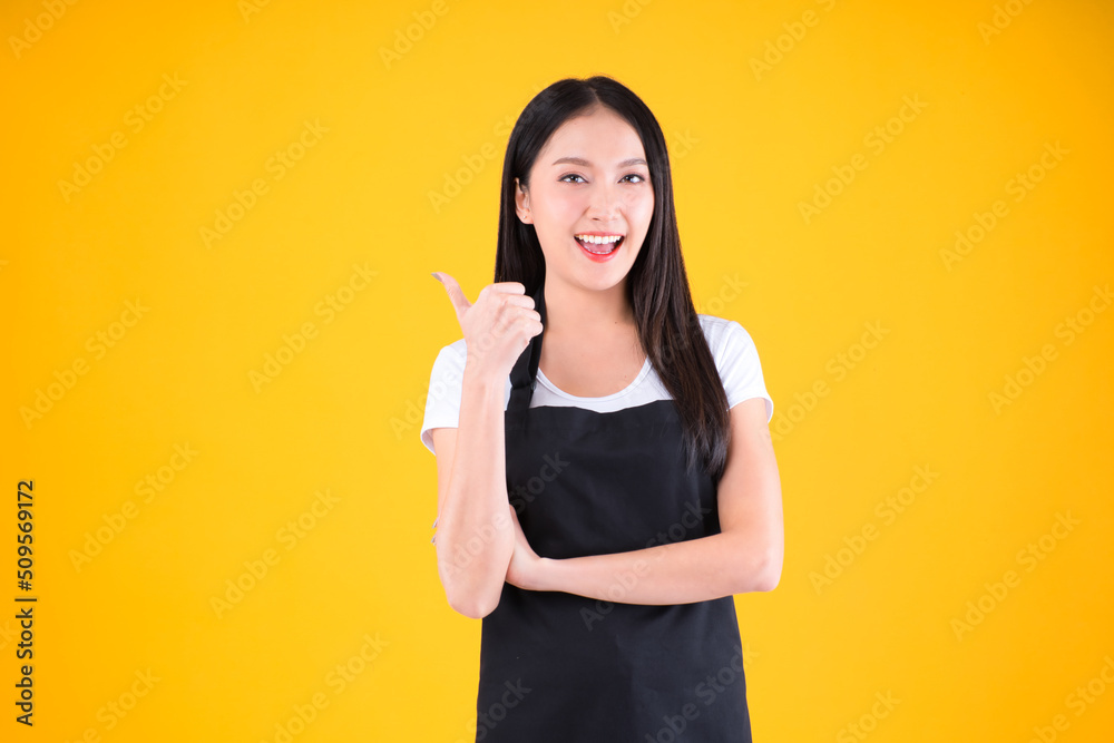 pretty asian female smile wearing black apron show and present pointing fingers copy space for your advertisement yellow background.